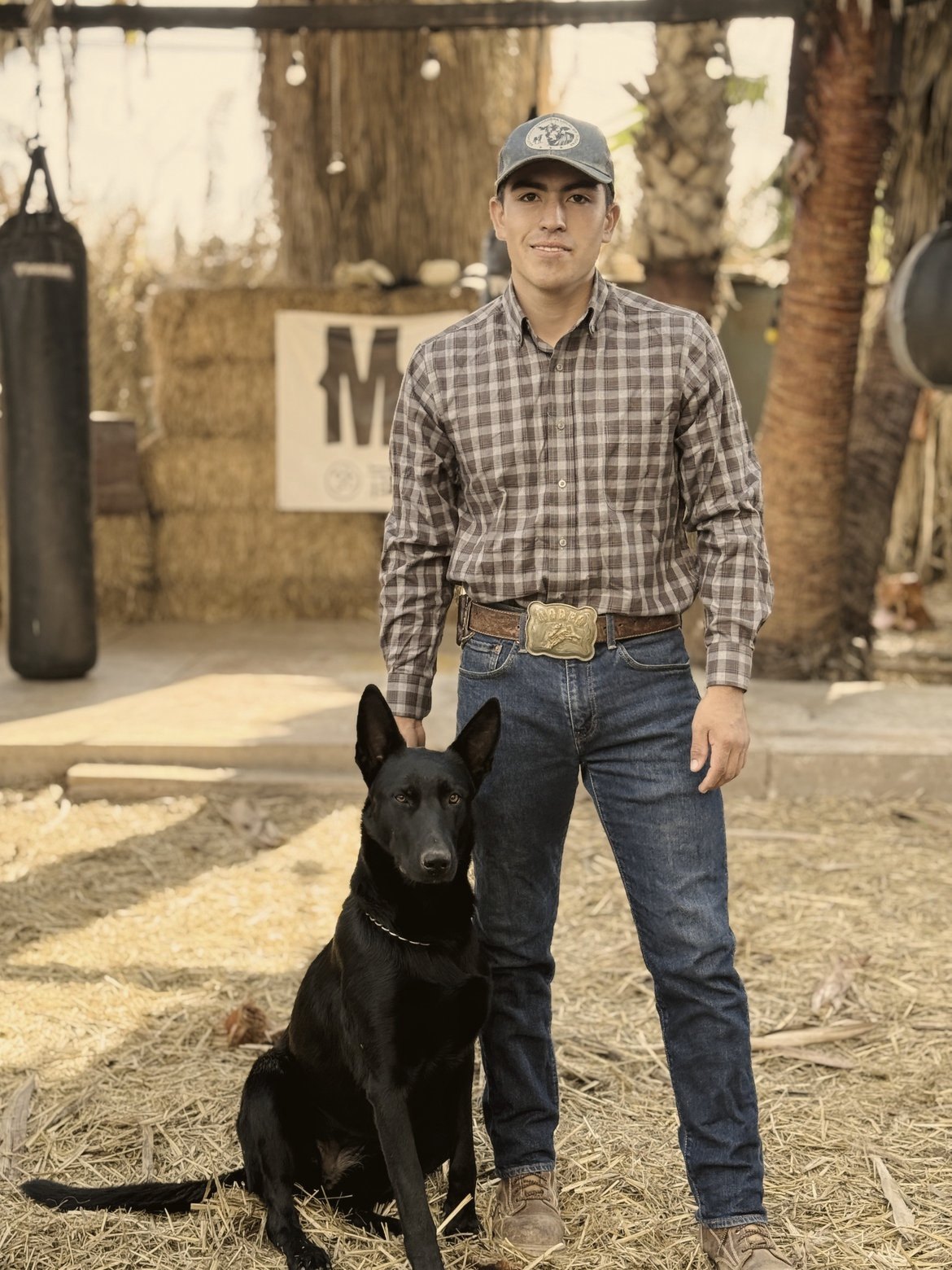 Abel Núñez Nuño, con camisa a cuadros, gorra, pantalón de mezclilla y hebilla de rodeo, de pie sobre paja junto a un perro pastor negro en el rancho de Mud & Lotus Baja