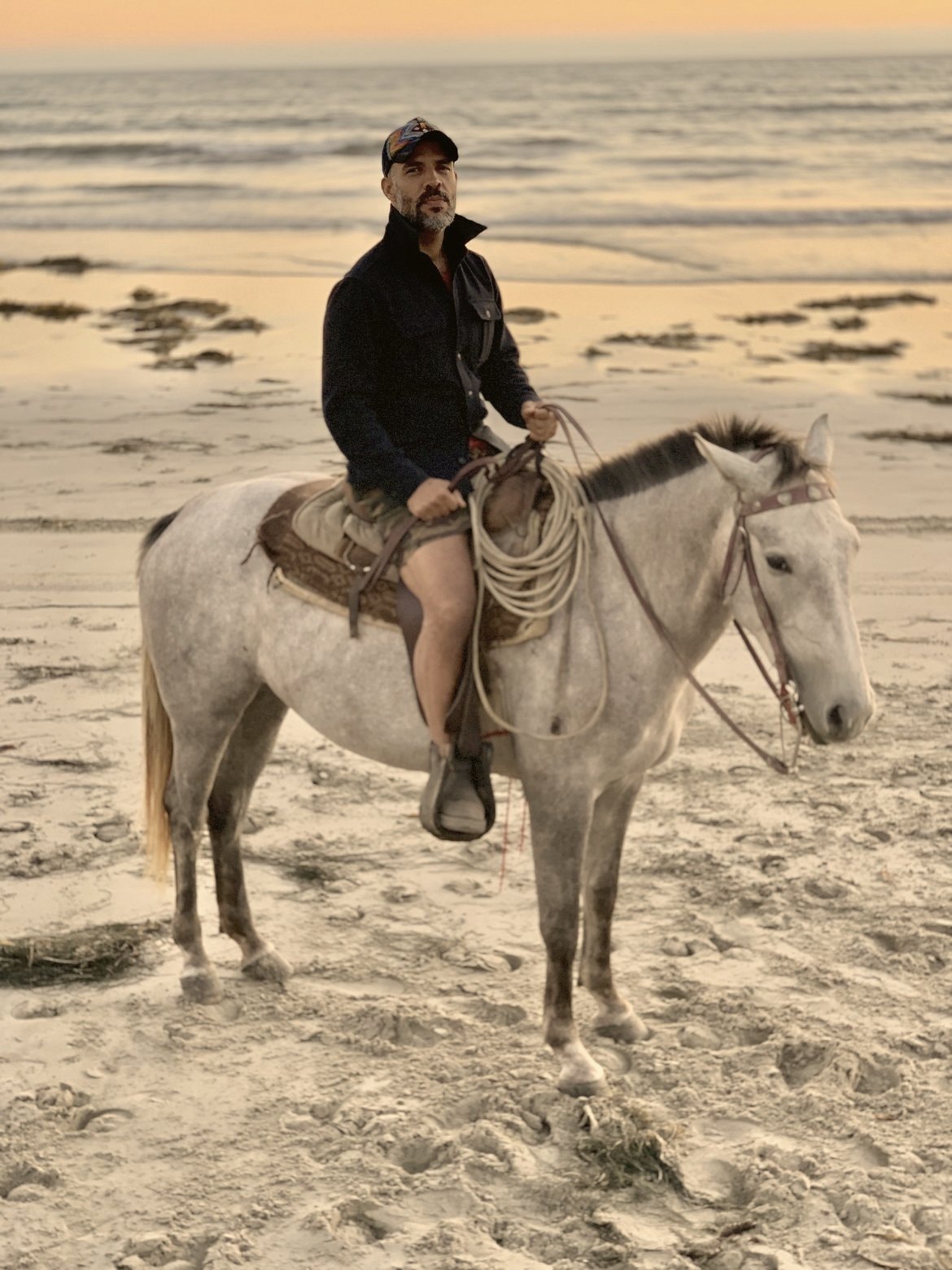 Anas “Coach” Canon, con chamarra oscura y gorra, montado en un caballo tordillo con una reata en la silla, en la playa al atardecer