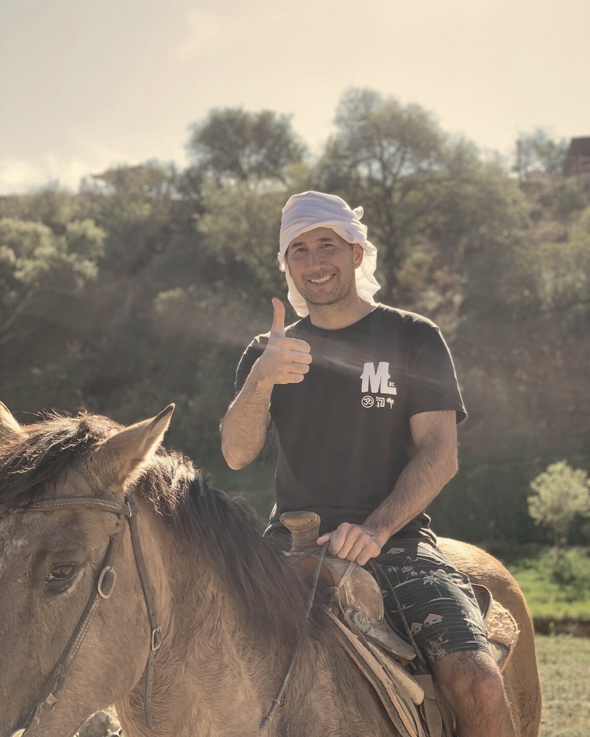 Un participante sonriente, con un pañuelo blanco en la cabeza, levanta el pulgar mientras monta un caballo café.
