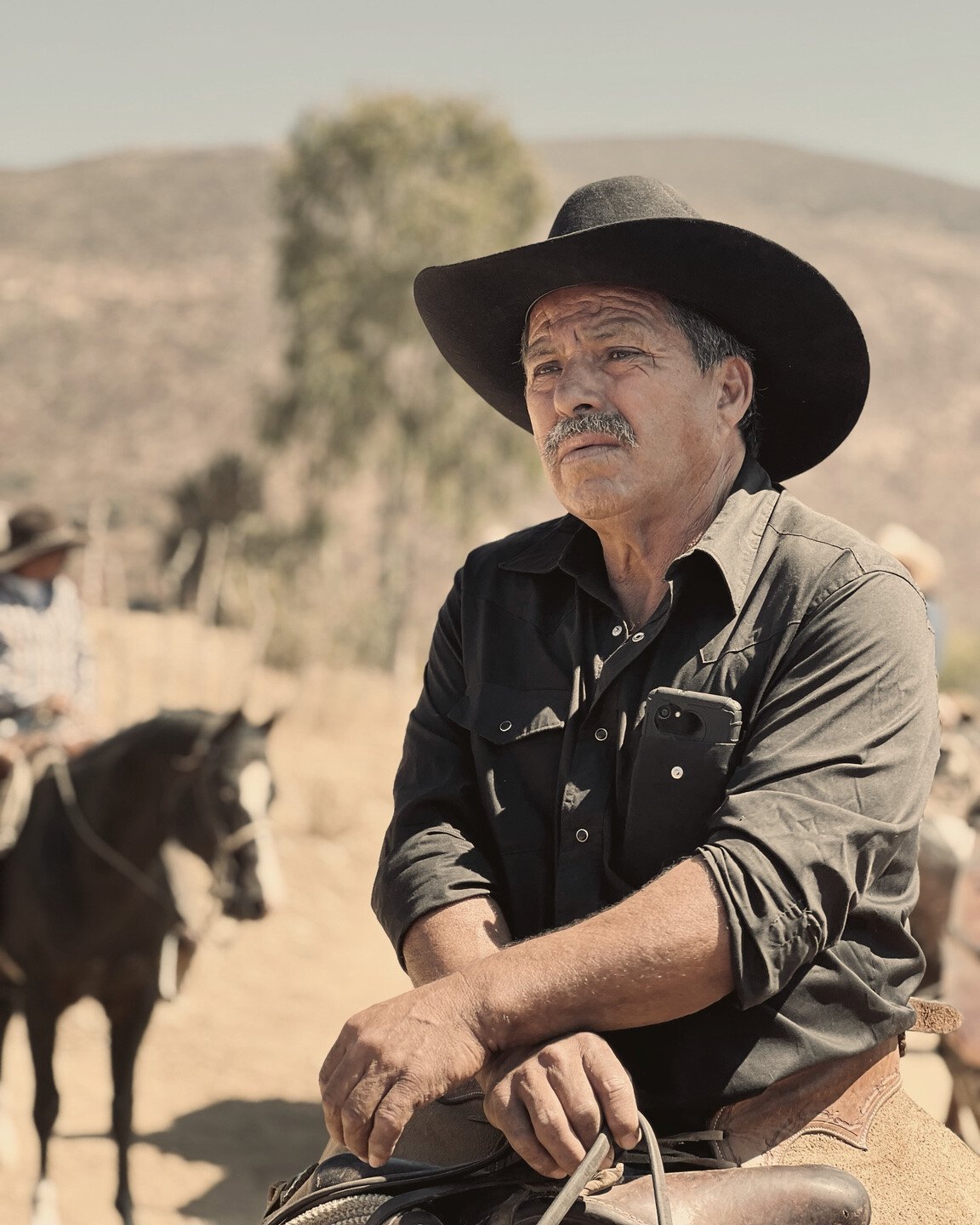 Un jinete con bigote, sombrero y camisa negros apoya los brazos sobre la montura, con otro caballo al fondo