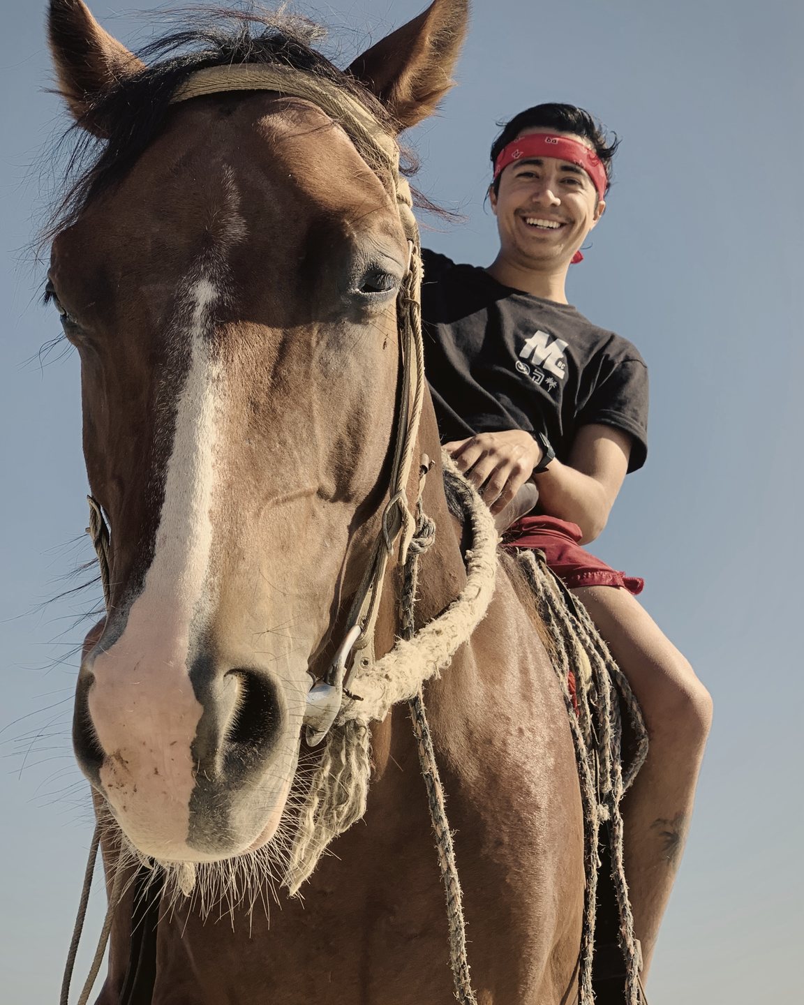 Un jinete sonriente con paliacate rojo, visto desde abajo junto a la cara de su caballo alazán, contra el cielo azul.