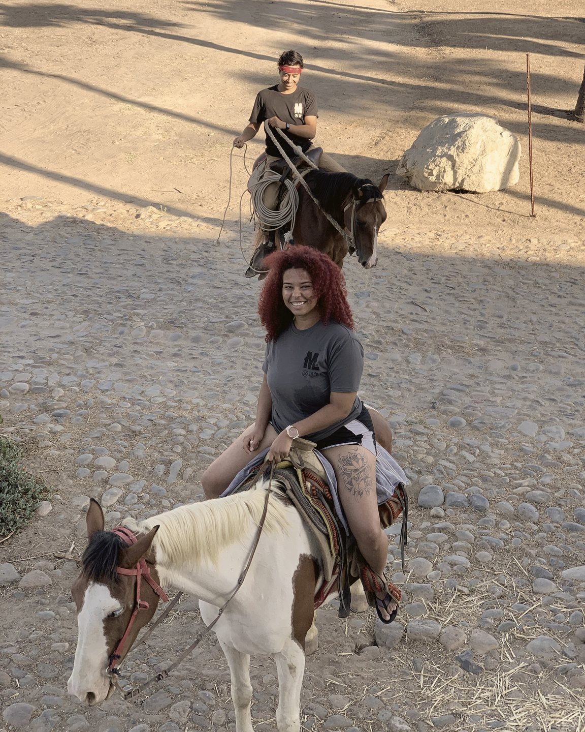 Una jinete sonriente de cabello rizado rojo en un caballo pinto, con otro jinete enrollando una reata detrás de ella.